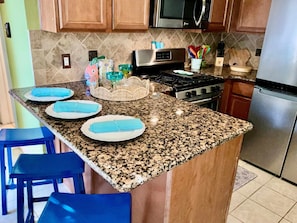 A kitchen with granite countertops, wooden cabinets, a stainless steel stove, and blue bar stools. - Surf & Strand Unit A for 5 (Galveston)