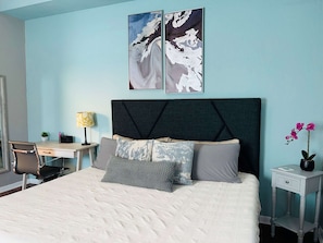 Un dormitorio ordenado con cama, escritorio, silla y mesita de noche con una orquídea en maceta.