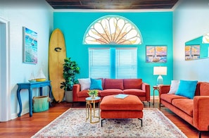 Una habitación con pared verde azulado, un ventanal con contraventanas, una tabla de surf, un sofá rojo, una mesita redonda y una planta en maceta.