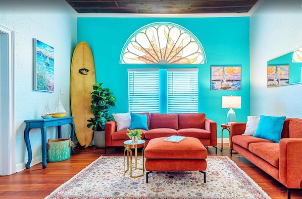 A room with a teal wall, a large window with shutters, a surfboard, a red sofa, a small round table, and a potted plant. - Surf & Strand Unit A for 5 (Galveston)