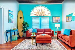 Una habitación con pared verde azulado, un ventanal con contraventanas, una tabla de surf, un sofá rojo, una mesita redonda y una planta en maceta.