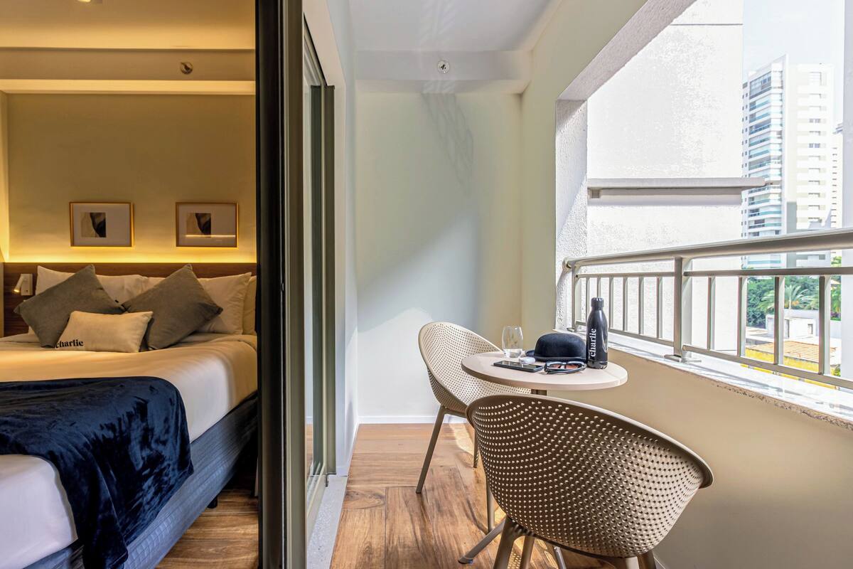Habitación de hotel con cama, zona de comedor con mesa y sillas, y vista de edificios a través de una ventana.