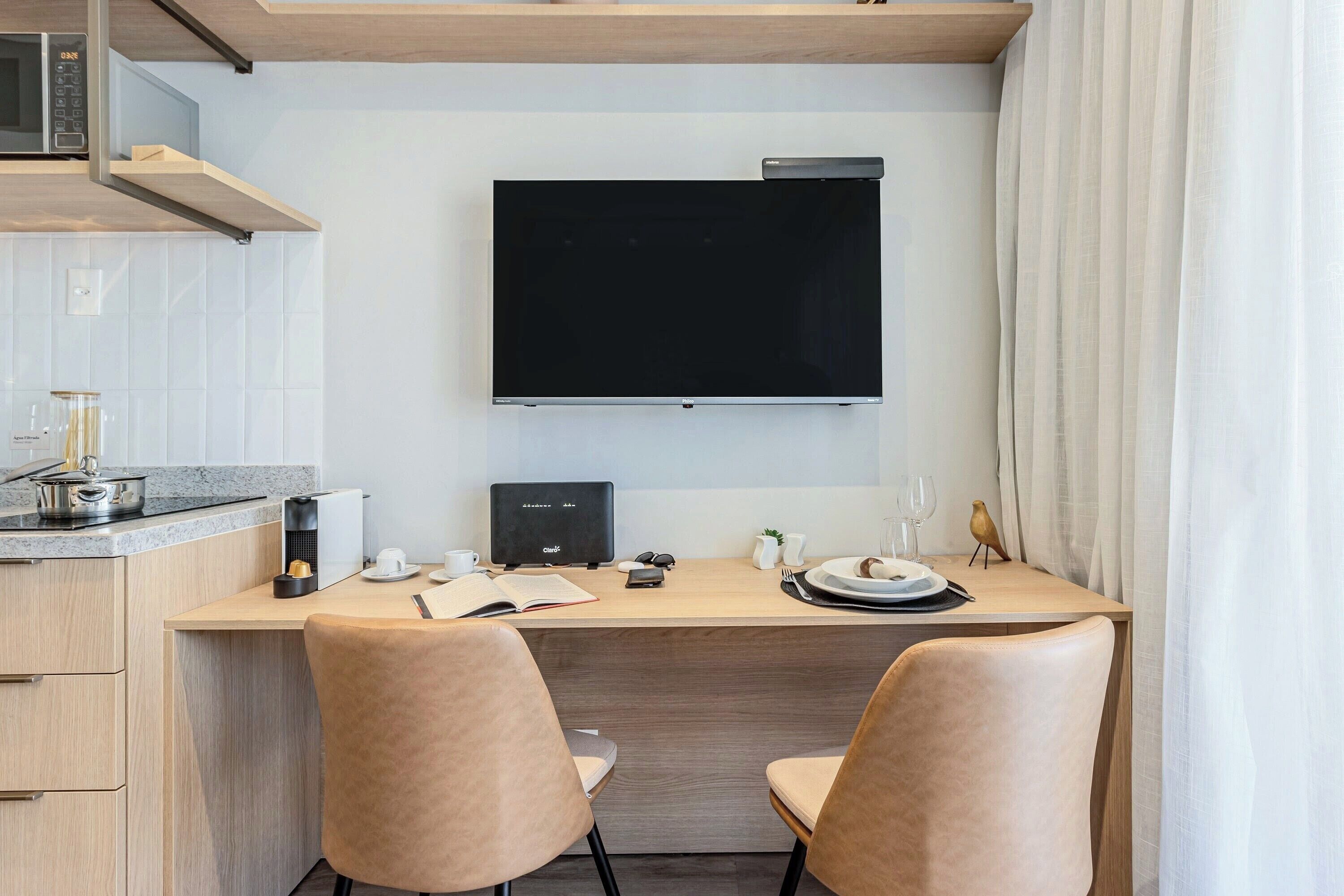 A modern kitchen with a flat-screen TV, wooden cabinets, a countertop with a kettle, and a dining area with two chairs.