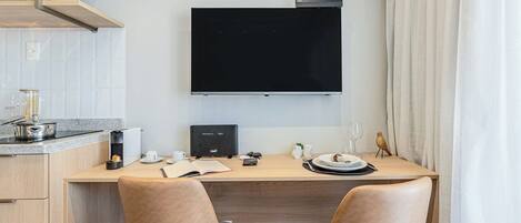 A modern kitchen with a flat-screen TV, wooden cabinets, a countertop with a kettle, and a dining area with two chairs.