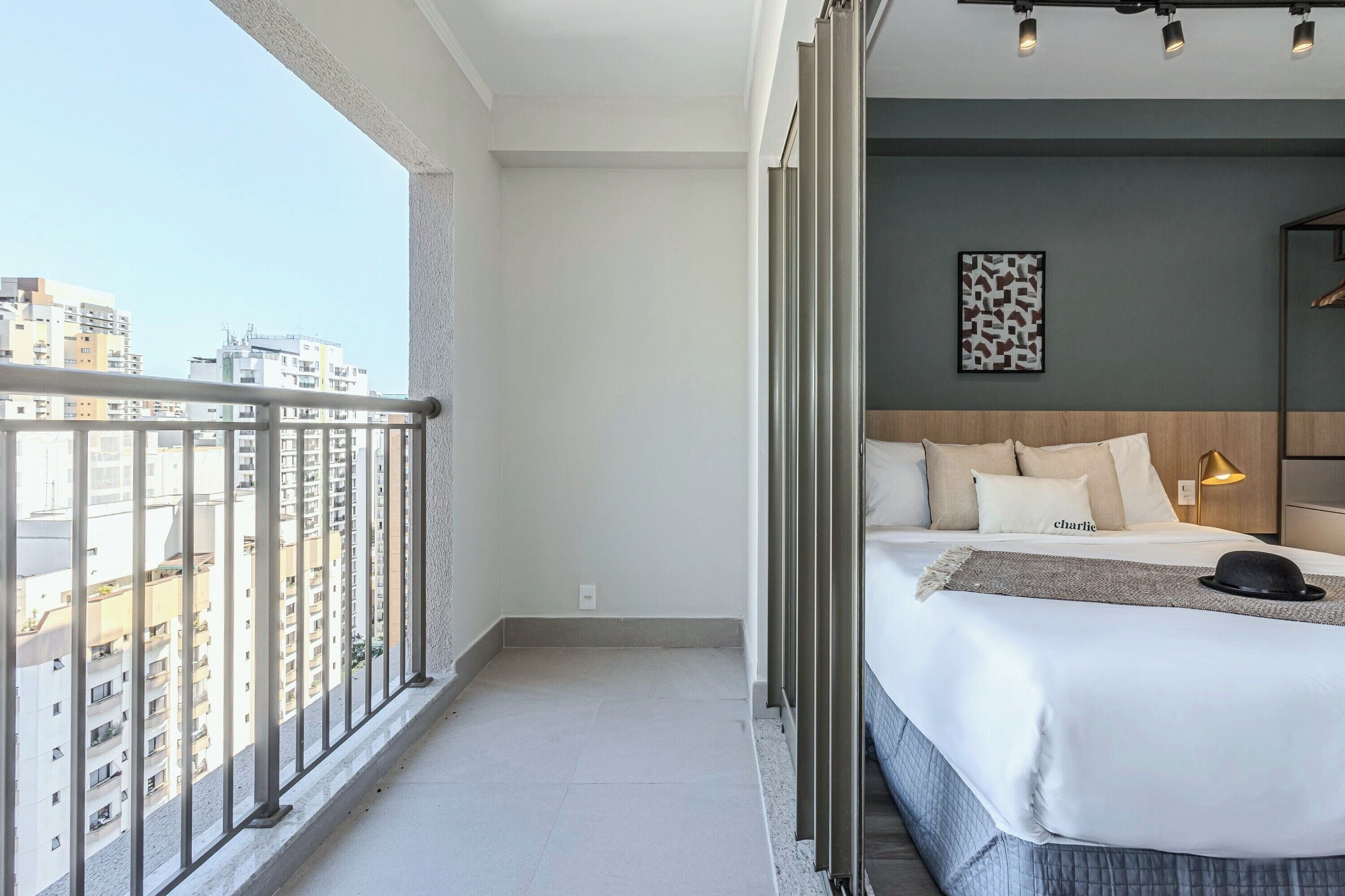 Une chambre d’hôtel avec un balcon, un lit avec une literie blanche, un chapeau noir posé sur le lit et une vue sur la ville.