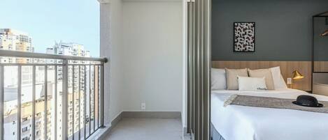 Une chambre d’hôtel avec un balcon, un lit avec une literie blanche, un chapeau noir posé sur le lit et une vue sur la ville.