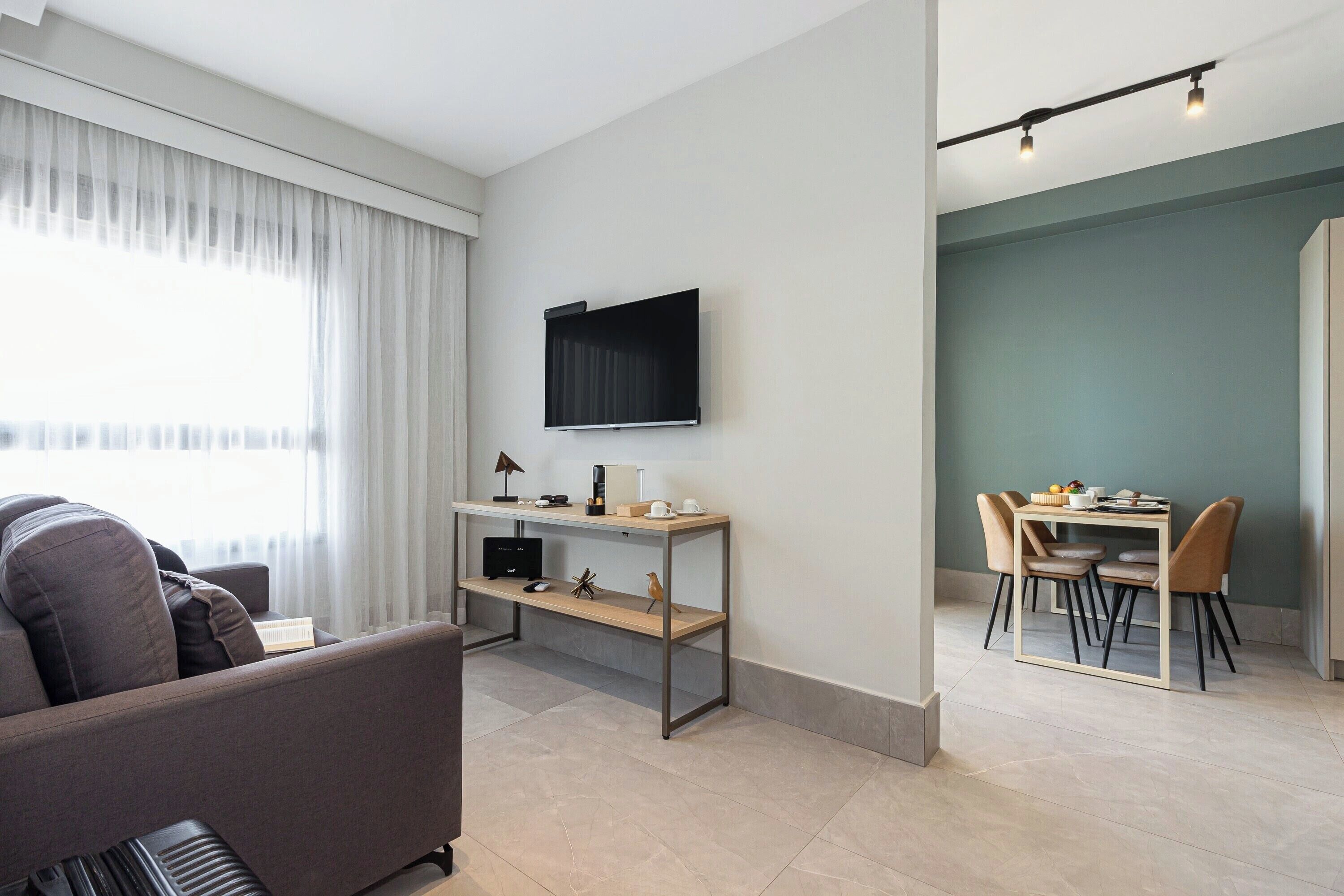 A modern living room with a sofa, a coffee table, and a TV mounted on the wall. There is a dining area with a table and chairs.