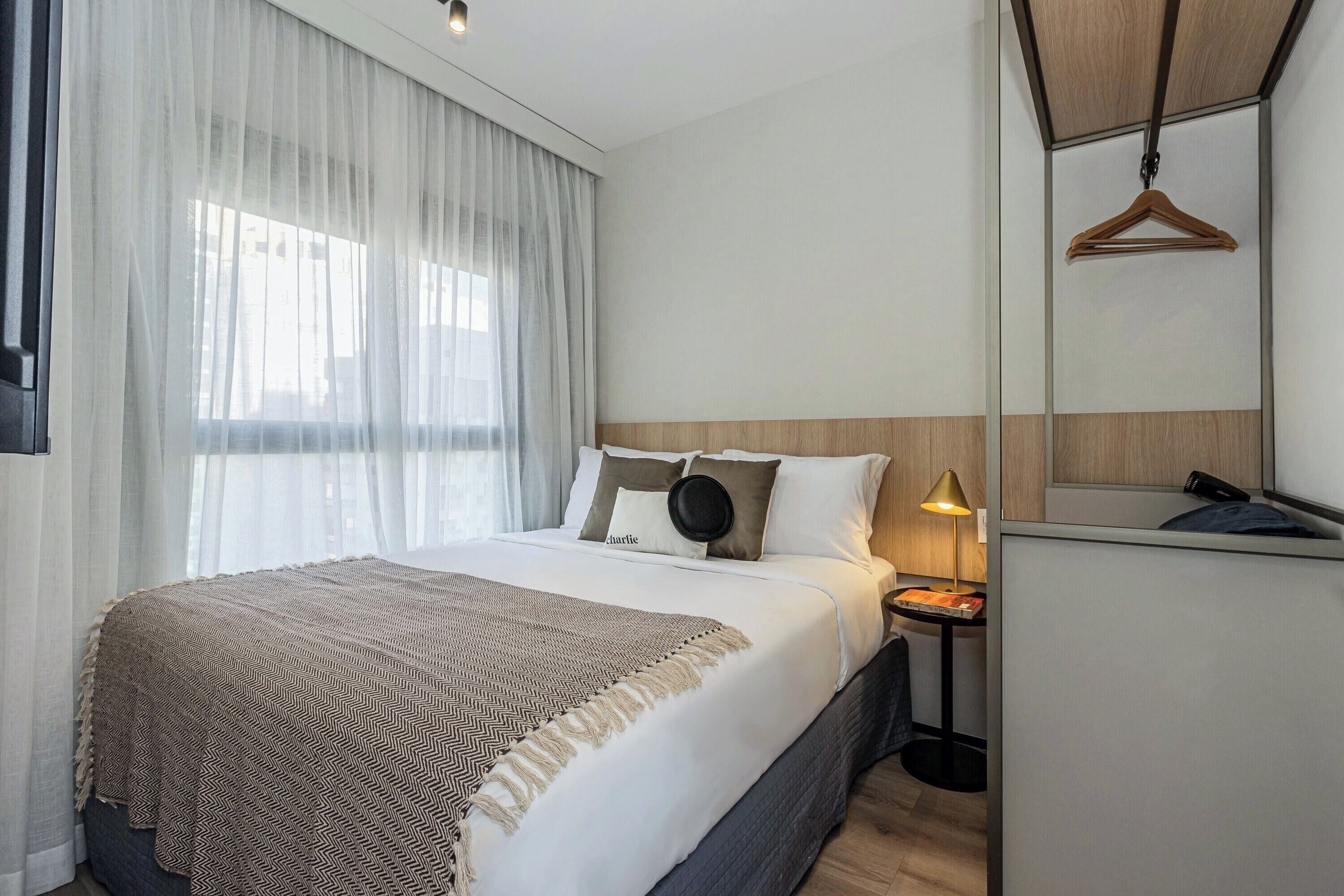 Une chambre d’hôtel avec un lit, une table de chevet avec une lampe, un porte-manteau mural et une vue sur l’extérieur à travers des rideaux transparents.