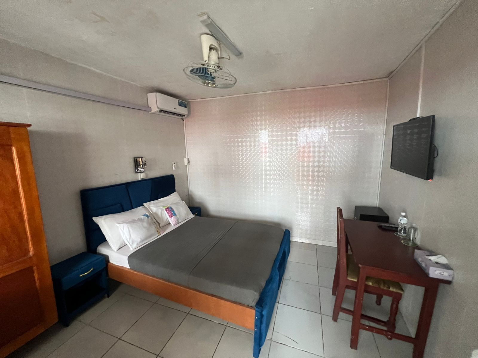 A hotel room with a bed, bedside table, desk, chair, and an air conditioning unit.