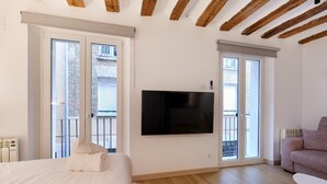 A modern bedroom with a large bed, a flat-screen TV mounted on the wall, and a balcony with a view of a brick building. - Apartamentos Linda by Clabao (Pamplona)