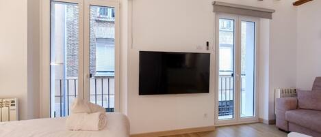 A modern bedroom with a large bed, a flat-screen TV mounted on the wall, and a balcony with a view of a brick building.