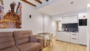 A modern living room with a sofa, a dining table with chairs, a kitchen area with white cabinets and appliances, and a ceiling with exposed beams. - Apartamentos Linda by Clabao (Pamplona)