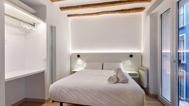 A modern bedroom with a large bed, wooden ceiling beams, a bedside table, and a view through a large window. - Apartamentos Linda by Clabao (Pamplona)