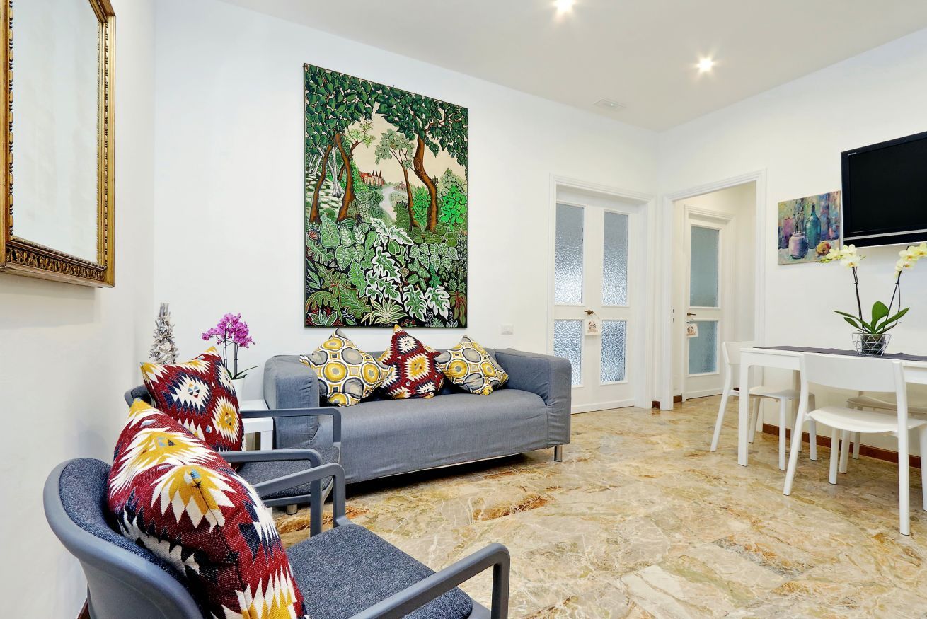 Een woonkamer met een grijze bank, gekleurde kussens, een schilderij van een bos, een eethoek met een witte tafel en stoelen, en een aan de muur bevestigde televisie.