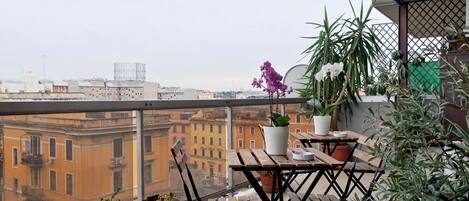 A balcony with a wooden table and chairs, potted plants, and a view of city buildings.