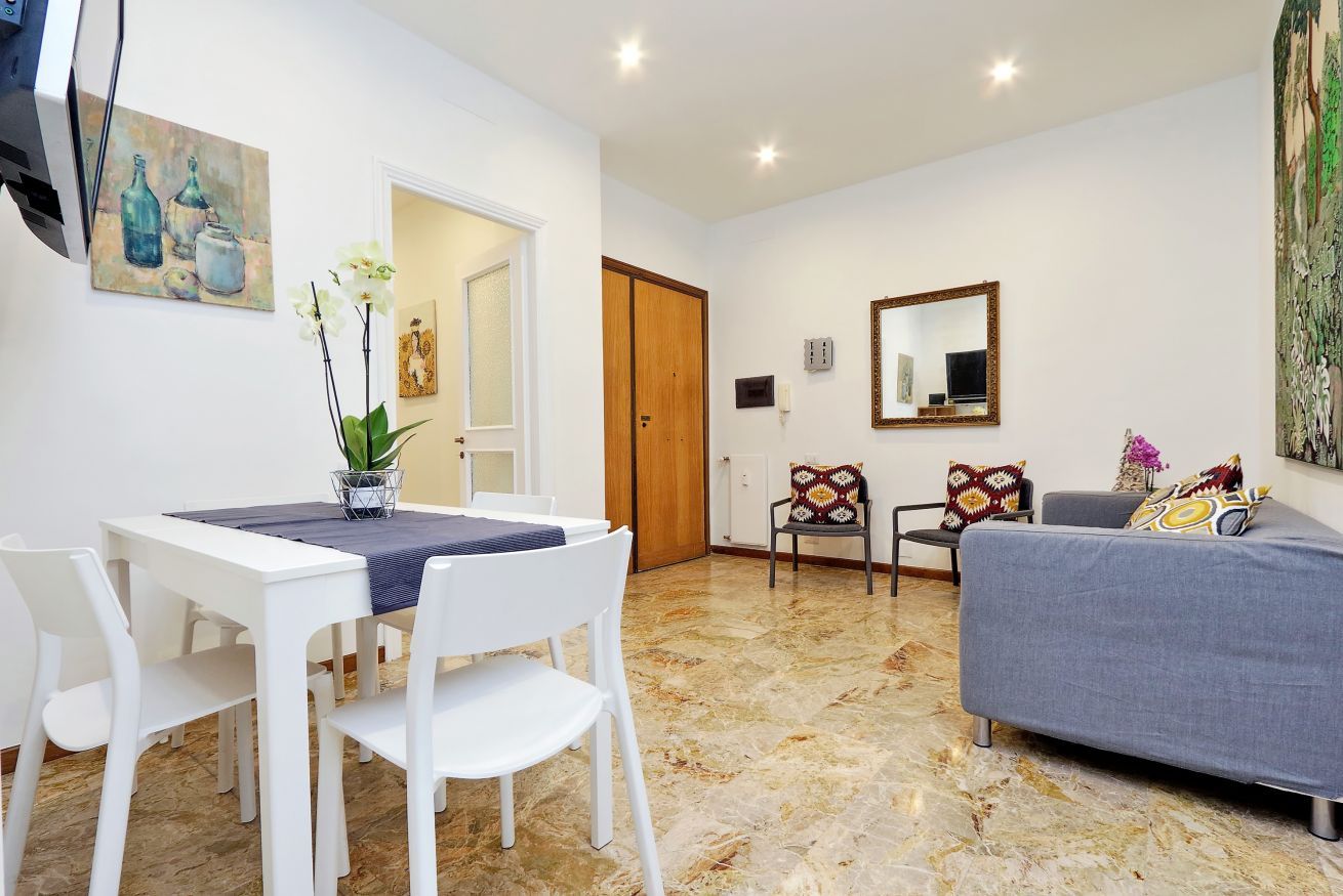 A modern living room with a dining table, white chairs, a grey sofa, and a patterned rug.