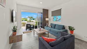 A modern living room with a grey sofa, a blue couch, and a wooden coffee table. There is a TV mounted on the wall, a potted plant, and a view of the outdoors through a sliding glass door. - Ocean Hibiscus Punta Azul Resort  (Sabana Westpunt)