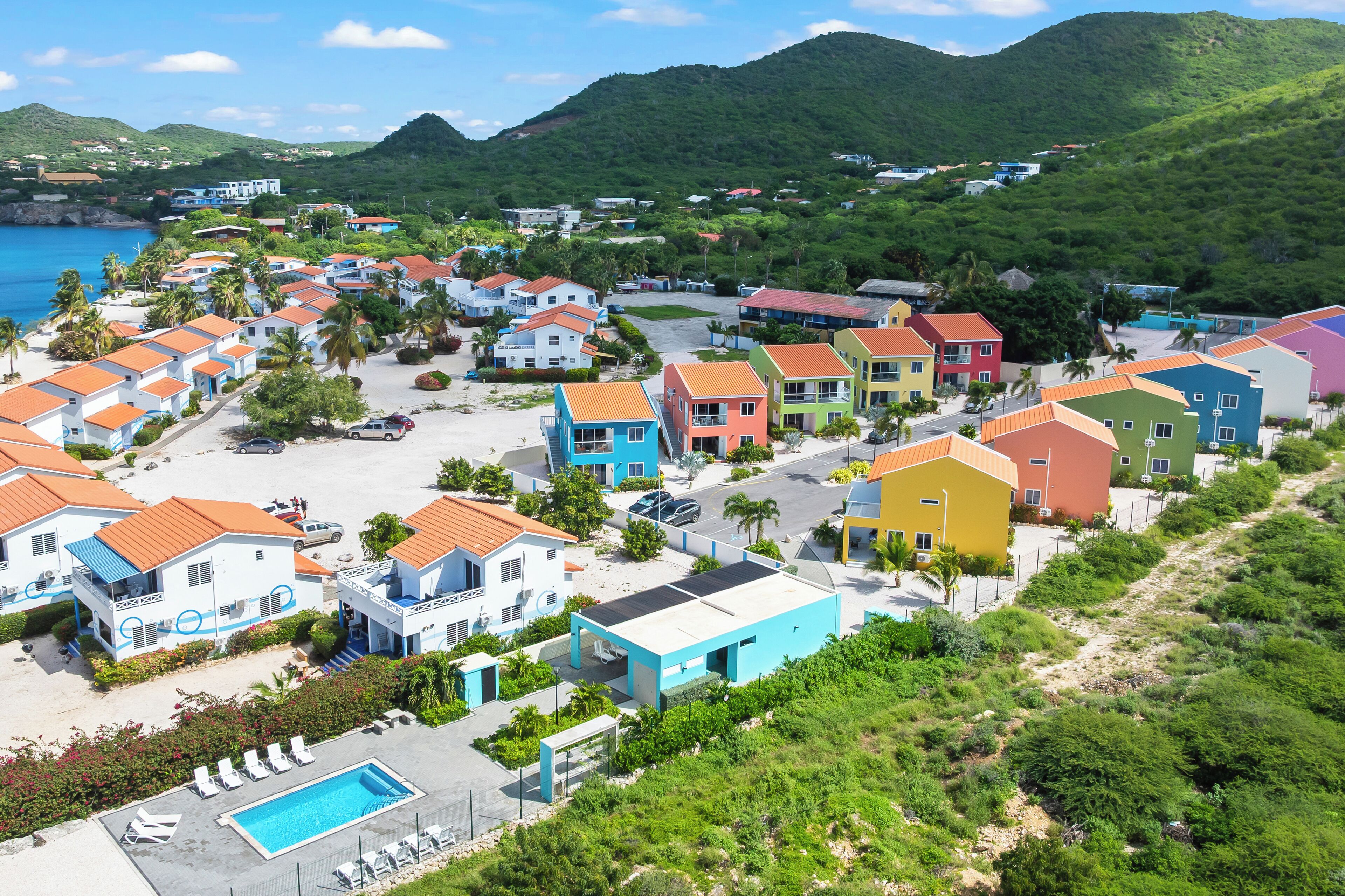 A coastal residential area with colorful houses, a swimming pool, and a hilly backdrop.
