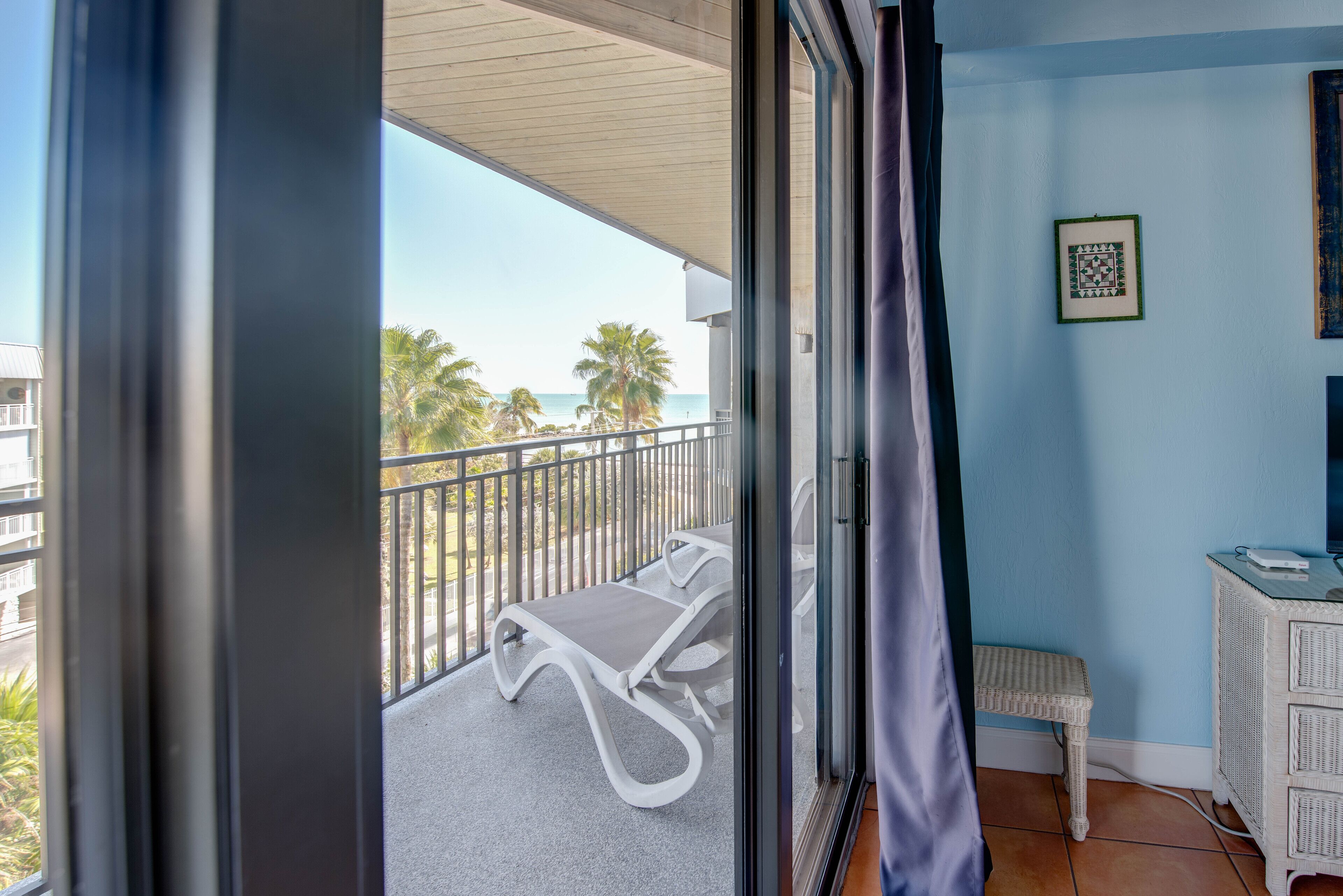 Een balkon met twee lounge fauteuils, een klein wit kattenhuis en uitzicht op het strand en palmbomen.