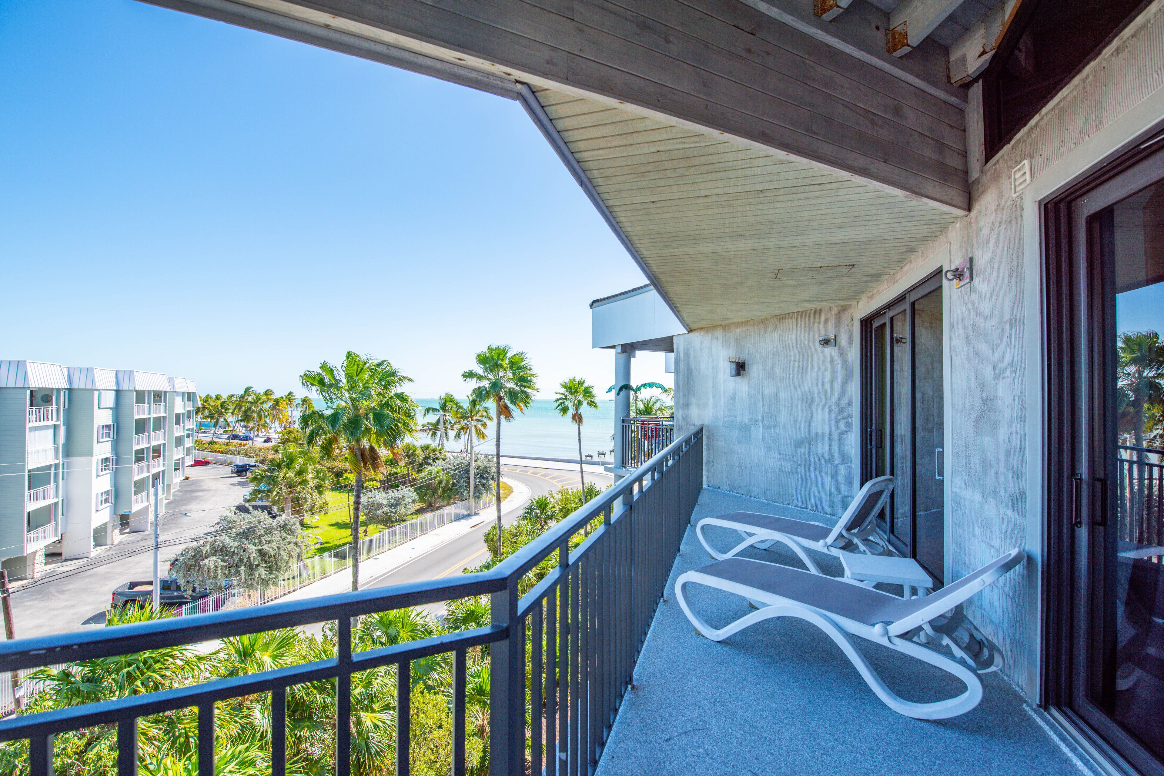 A balcony with a view of a road, palm trees, and a building across the street.