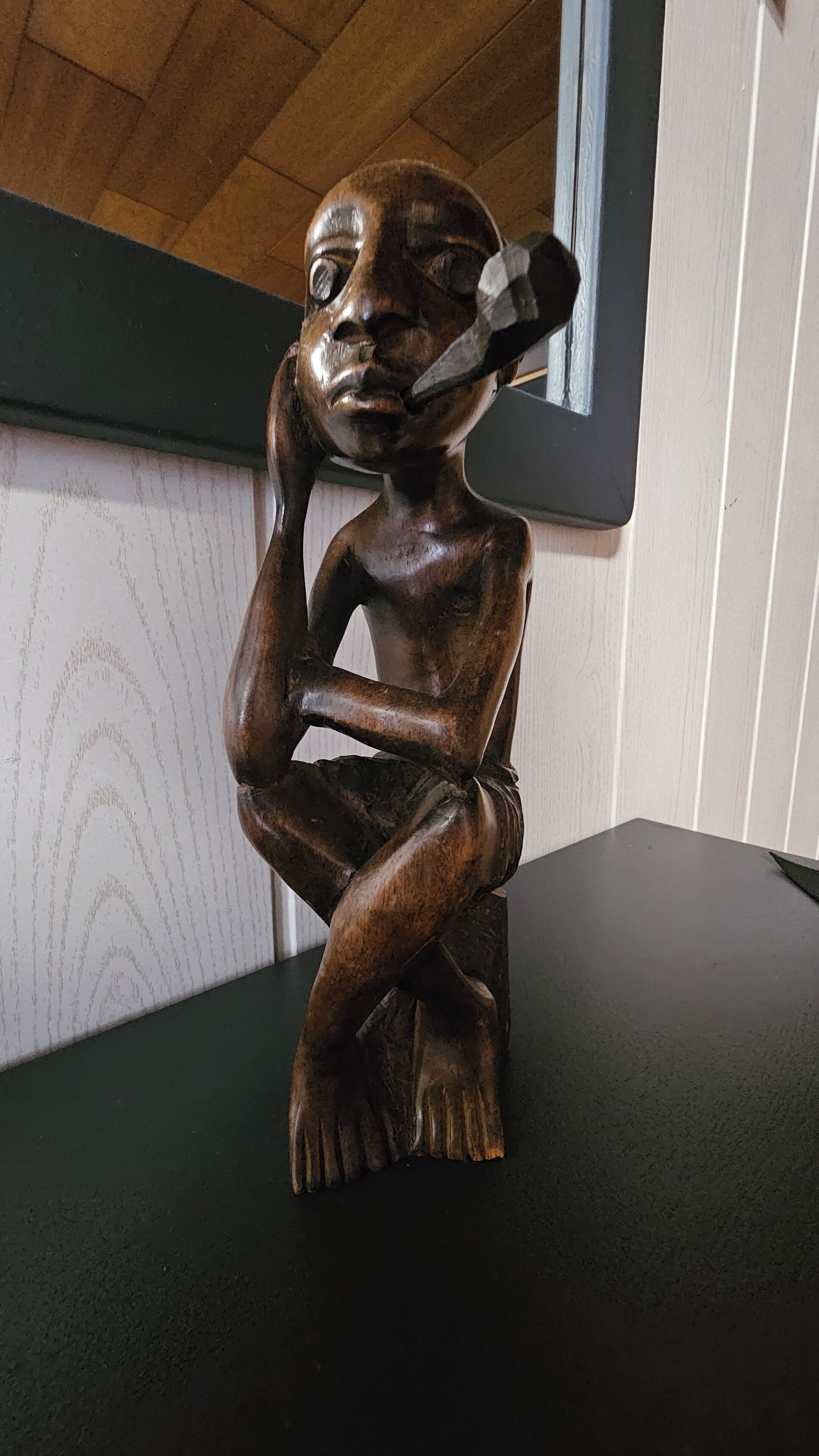 A wooden sculpture of a seated figure with a contemplative pose, placed on a dark surface against a wall with a patterned design.