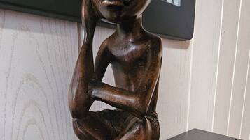 A wooden sculpture of a seated figure with a contemplative pose, placed on a dark surface against a wall with a patterned design.