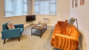 A living room with a blue armchair, an orange sofa, a coffee table, and a TV on the wall.