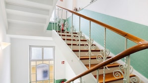 Una escalera con barandillas metálicas ornamentadas y pasamanos de madera, que conduce a una habitación con pared verde y ventana de vidrio satinado.