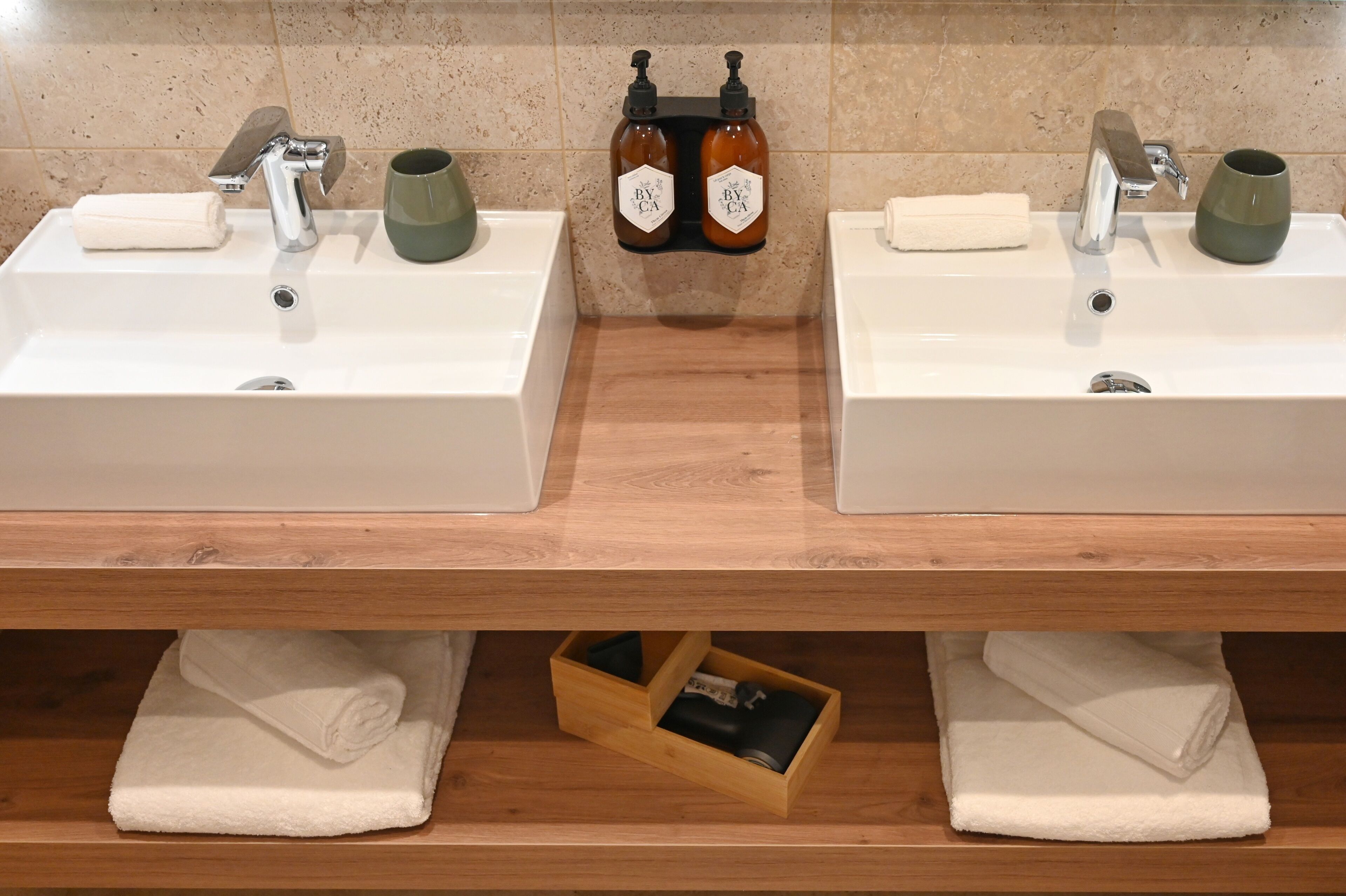 A bathroom with two sinks, each with a soap dispenser and a towel. There are two towels folded under the sinks and a wooden shelf with a box.