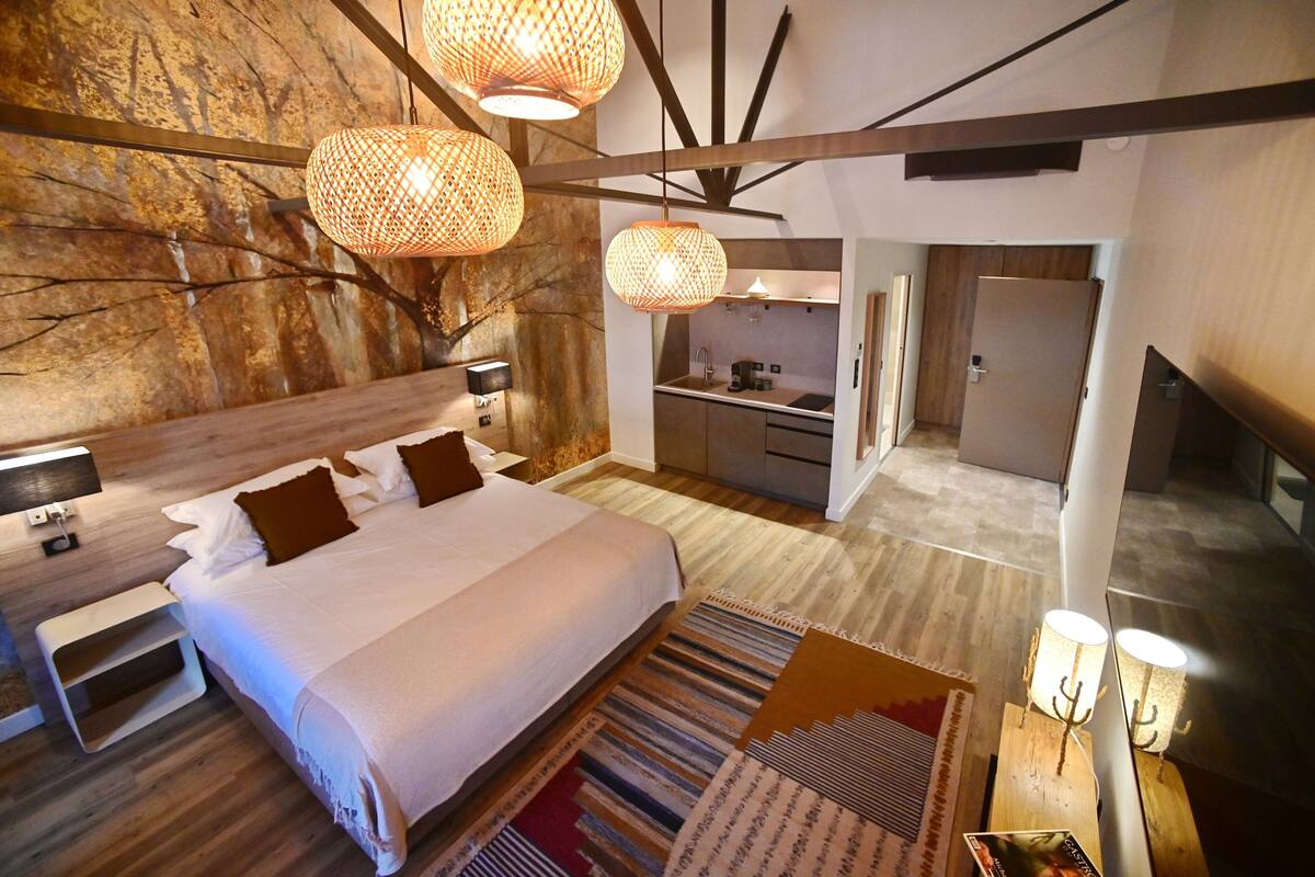 A modern hotel room with a large bed, bedside tables, a built-in kitchen, and a unique stone wall.