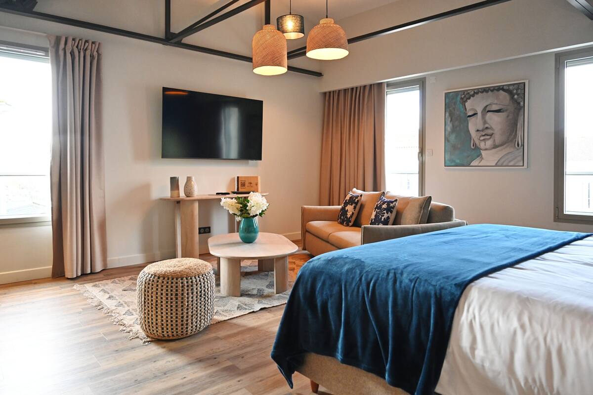 A modern hotel room with a large bed, a sofa, a small round table, and a flat-screen TV mounted on the wall.
