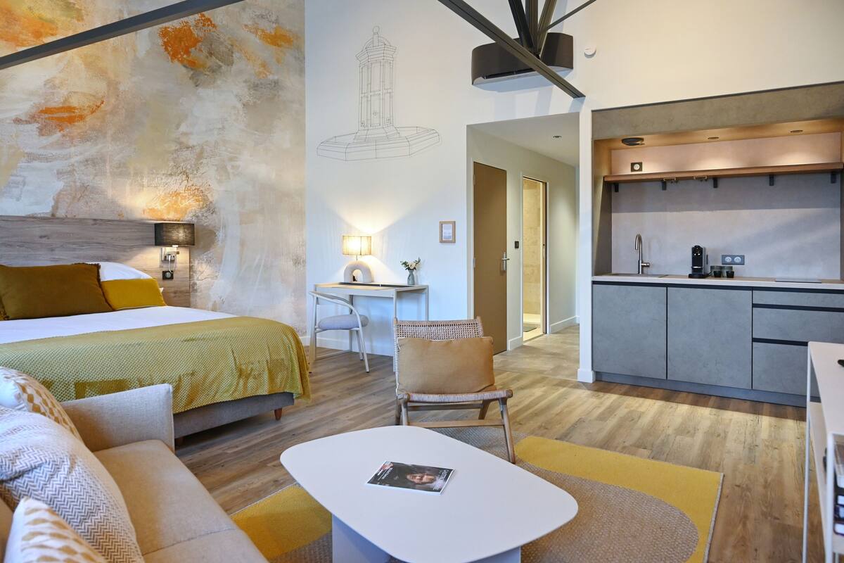 A modern hotel room with a bed, a desk, a chair, a small table, and a kitchenette.