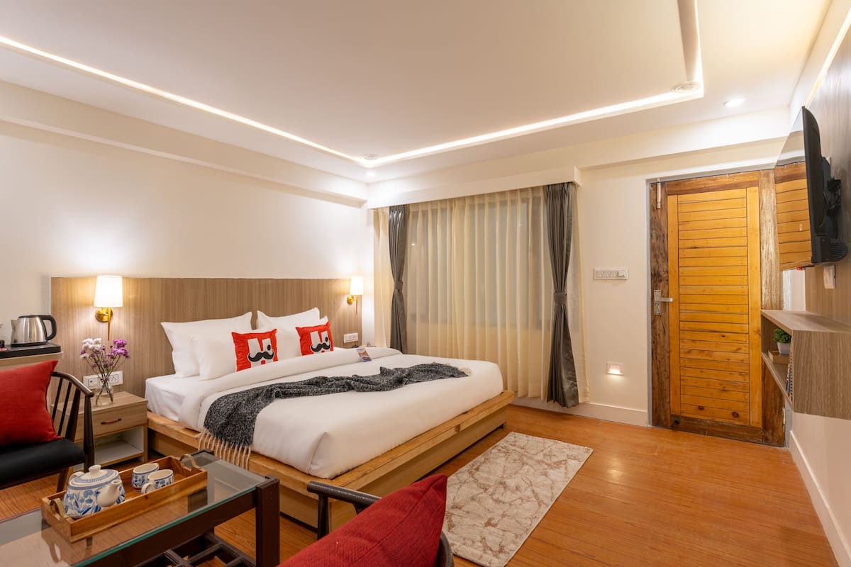 A hotel room with a bed, bedside table, lamp, curtains, wooden door, and a TV mounted on the wall.