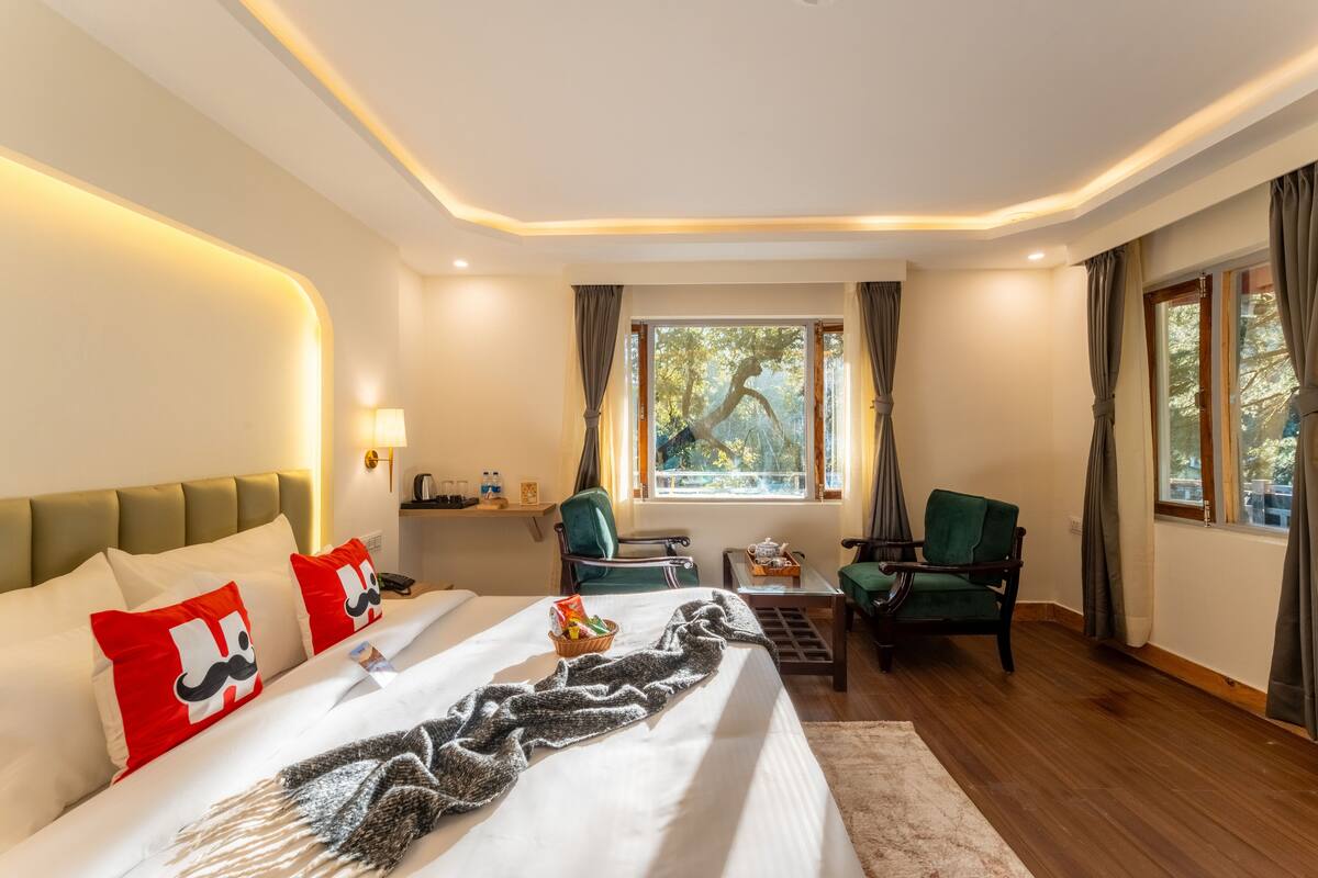 A hotel room with a large bed, two green armchairs, a small table with a teapot, and a window with curtains.