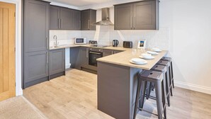 A modern kitchen with grey cabinets, a central island with stools, a microwave, and a kettle.