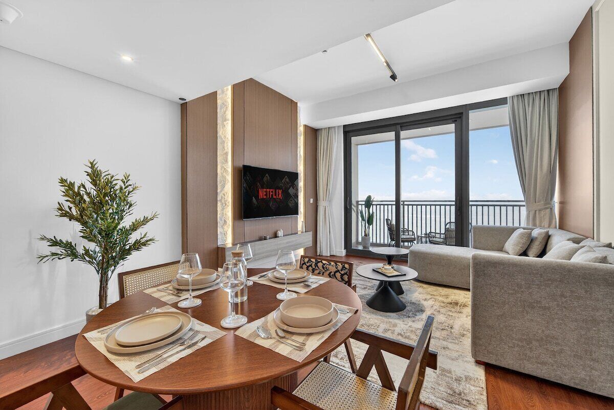 A modern living room with a dining area, a flat-screen TV mounted on the wall, and a balcony with a view.