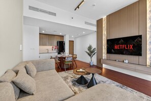 Un salon moderne avec un canapé d’angle, un coin repas avec une table et des chaises, une cuisine en arrière-plan et un téléviseur fixé au mur qui affiche le logo de Netflix.