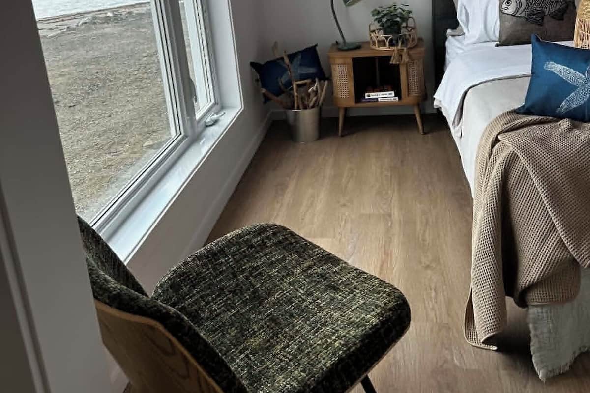 A bedroom with a large window, a bed with a headboard, a bedside table with a lamp, and a chair.