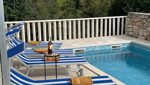 Ein Poolbereich mit blau gestreiften Liegestühlen, einem kleinen Tisch mit Flasche und Glas sowie einer Steinwand im Hintergrund.