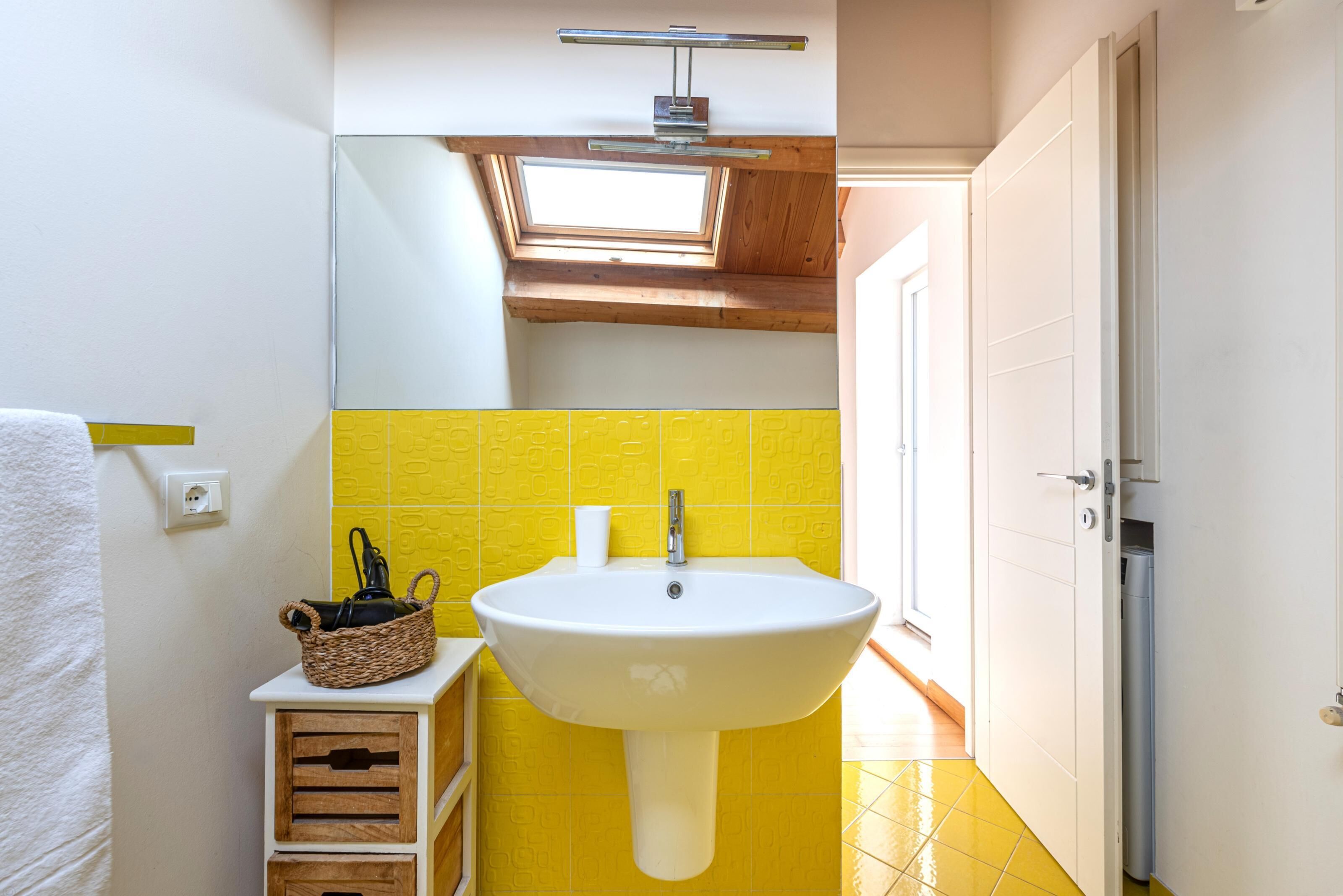 Un baño con un lavamanos blanco, azulejos amarillos, un tragaluz y una cesta sobre un mueble de madera.