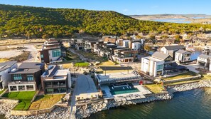 A modern residential area with contemporary houses, a central pool, and a view of a lake and forested hills. - Lake Luxury- Juniper Ridge-Pool Table and Beautiful Decor (Graford)