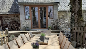 Une terrasse en bois avec une longue table et des chaises, des plantes en pot et, au fond, un bâtiment en pierre.