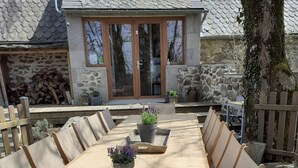Een houten terras met een lange tafel en stoelen, plantenbakken en een stenen gebouw op de achtergrond.