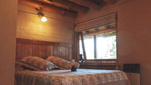 Une chambre avec une tête de lit en bois, un lit recouvert d’une literie à motifs, une petite table sur laquelle est posé un vase, et une fenêtre donnant sur un paysage verdoyant.