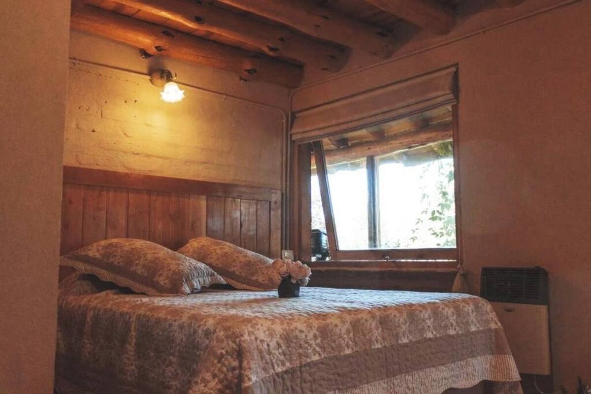 Un dormitorio con cabecera de madera, cama con ropa de cama estampada, una mesita con un florero y una ventana con vista a follaje.