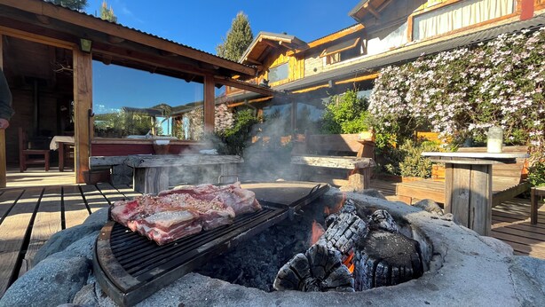 Un barbecue avec de la viande, un foyer en pierre et une terrasse en bois avec du mobilier d’extérieur.
