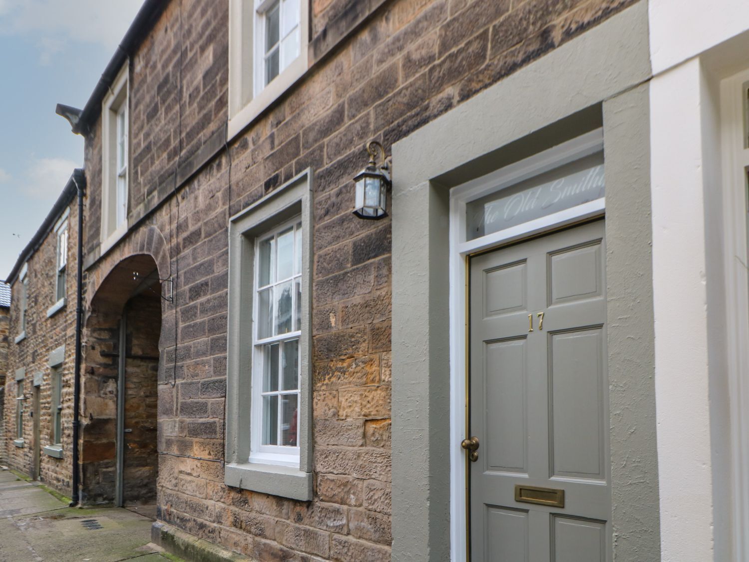 A row of stone buildings with a central arched doorway, a door with a number plaque, and a small window with a light fixture.