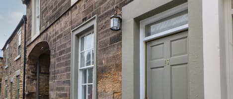 A row of stone buildings with a central arched doorway, a door with a number plaque, and a small window with a light fixture.