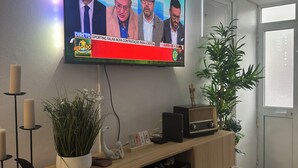 A television mounted on a wall displaying a news broadcast. - Casa Santa Maria (Vila do Porto)