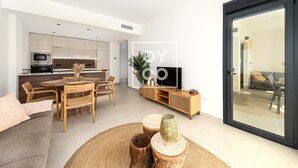 A modern living room with a sofa, dining table, and chairs, a kitchen area with appliances, and a sliding glass door leading to an outdoor area. - Amalia Garden Groundfloor Retreat, a footstep from beach and sea, By OYOOSTAYS (Torrevieja)